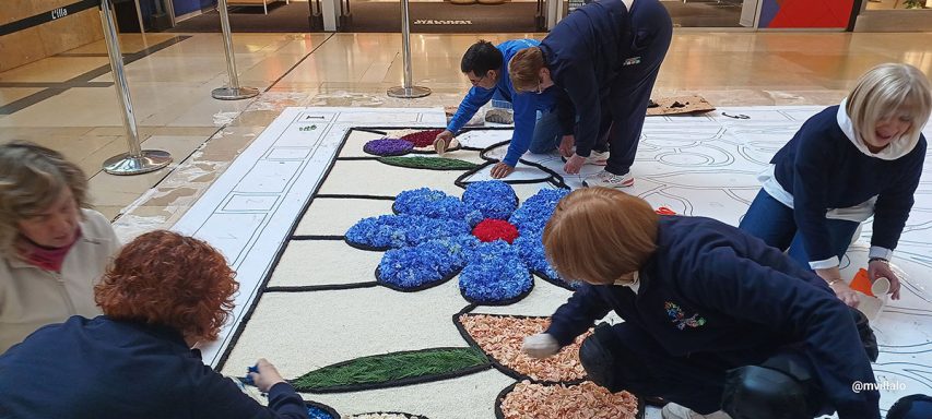 Varias personas trabajando en un gran tapiz de flores en un espacio abierto.